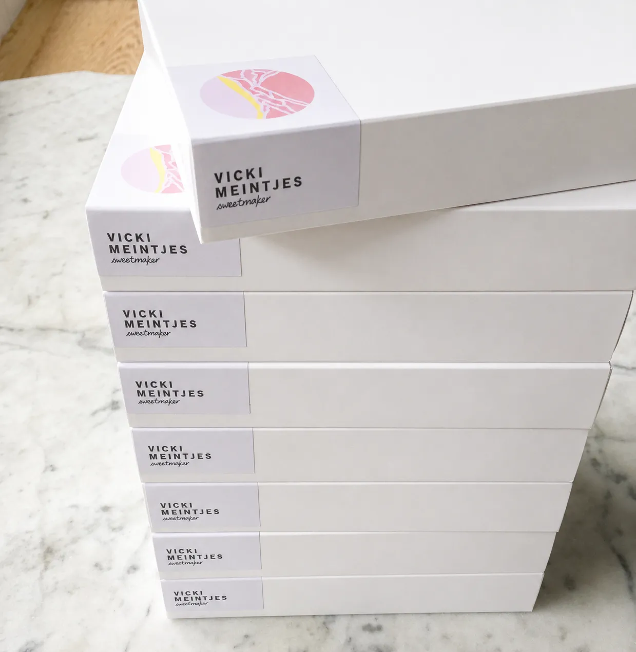 A stack of Vicki Meintjes Sweet Maker cake boxes, each finished with the brand mark sticker on the end panel.