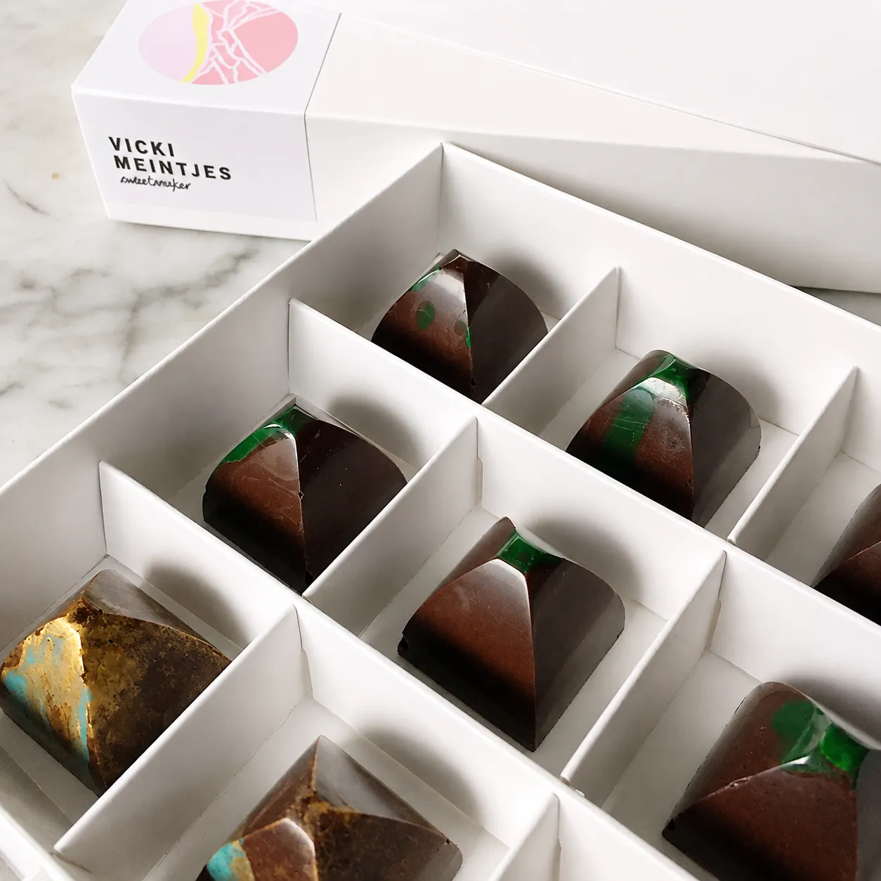 An open box of Vicki Meintjes chocolates with the fractured brand mark sticker and wordmark on the lid.
